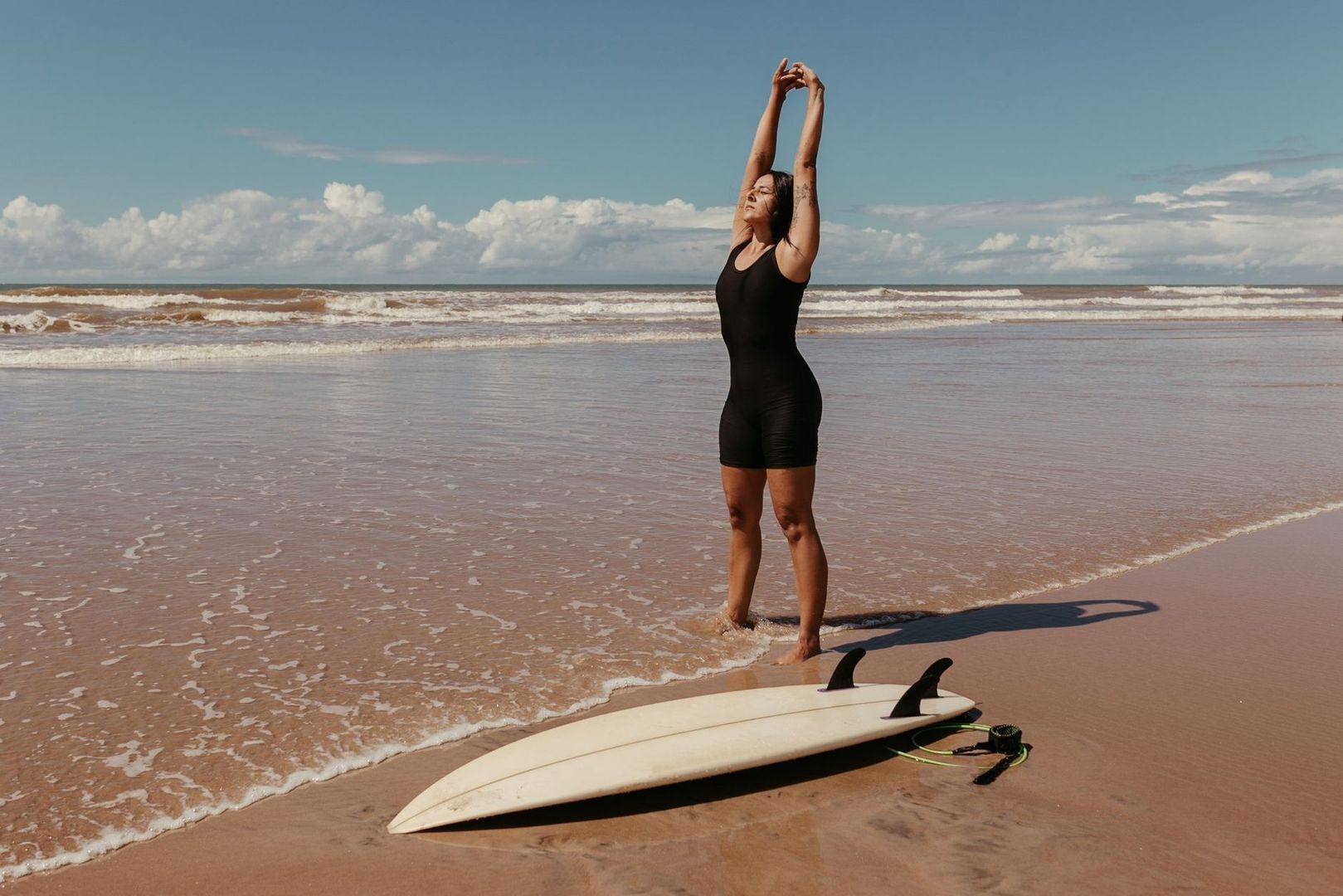 Surfer stretching in shallow water beside a surfboard on the beach