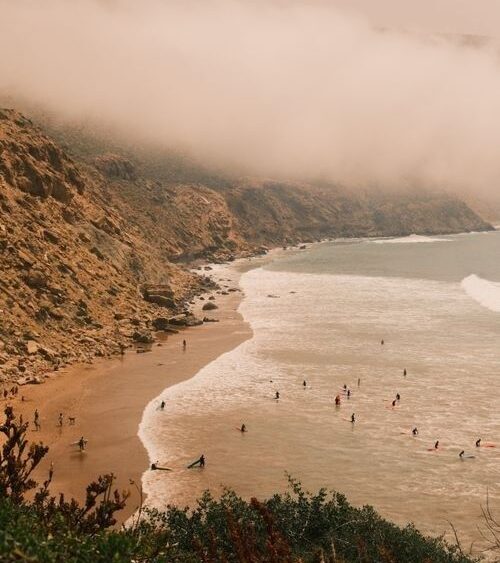 Foggy coastal beach with swimmers, rocky cliffs, and gentle waves
